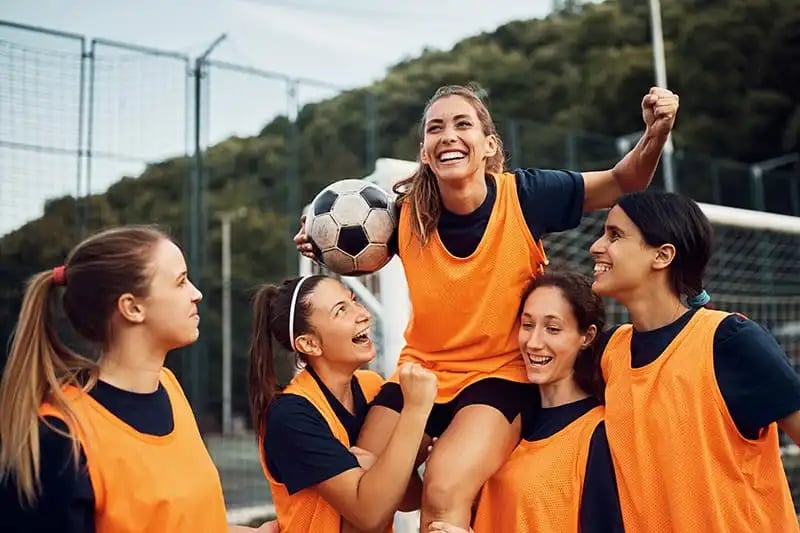 A team of women playing soccer and raising a teammate in the air representing their latest win.